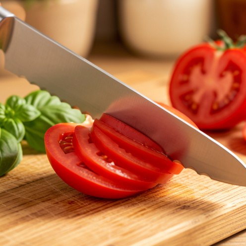 Couteau à tomate dentelé tranchant une tomate rouge mûre en fines rondelles régulières sur une planche en bois.